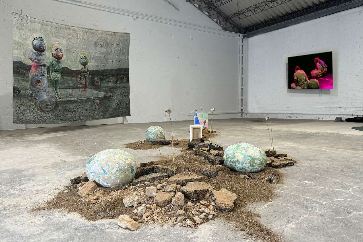 A Growing Wave Breaks in the Shore of Time — glazed ceramic poppy heads emerging from excavated soil and asphalt rubble in a hangar-like hall, with a Jacquard tapestry hung behind.