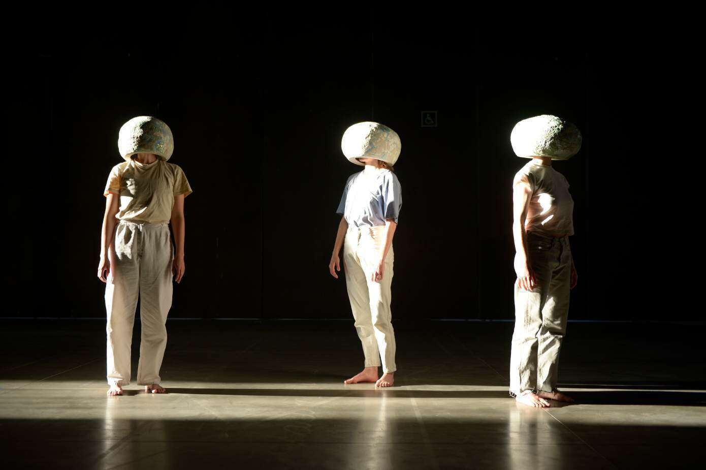 The Dance of the Opium Poppies — three performers in pale clothing stand on a dark stage, each wearing a large ceramic poppy-head over the head.
