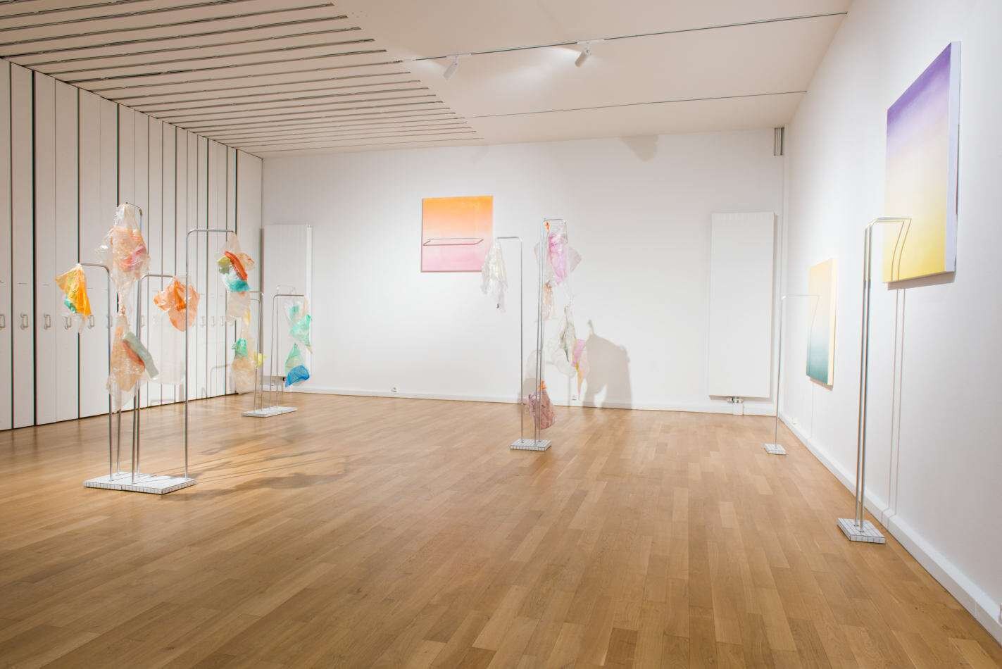 Toxic Secrets (Oikos) — translucent dyed plastic forms suspended on slim metal stands across a gallery, with gradient panels on the walls.